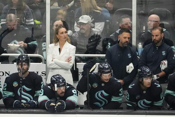 A white woman with blo<em></em>nde hair in a white suit stands behind a row of hockey players seated on a bench, with her arms crossed.