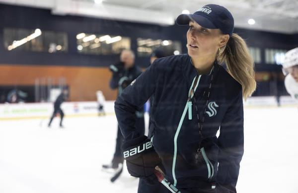 A white woman with blo<em></em>nde hair in a ponytail, wearing a ba<em></em>seball cap and a zip-up jacket, stands on a skating rink holding a hockey stick.