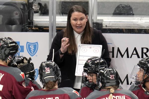 A white woman with long, brown hair holds up a whiteboard with a hockey rink diagram on it while speaking to a crowd of hockey players