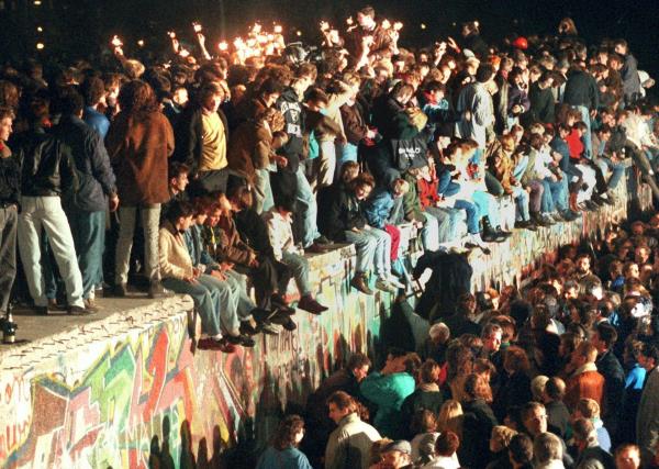 A historical photo of people standing and sitting on the Berlin wall as it is a<em></em>bout to be torn down.